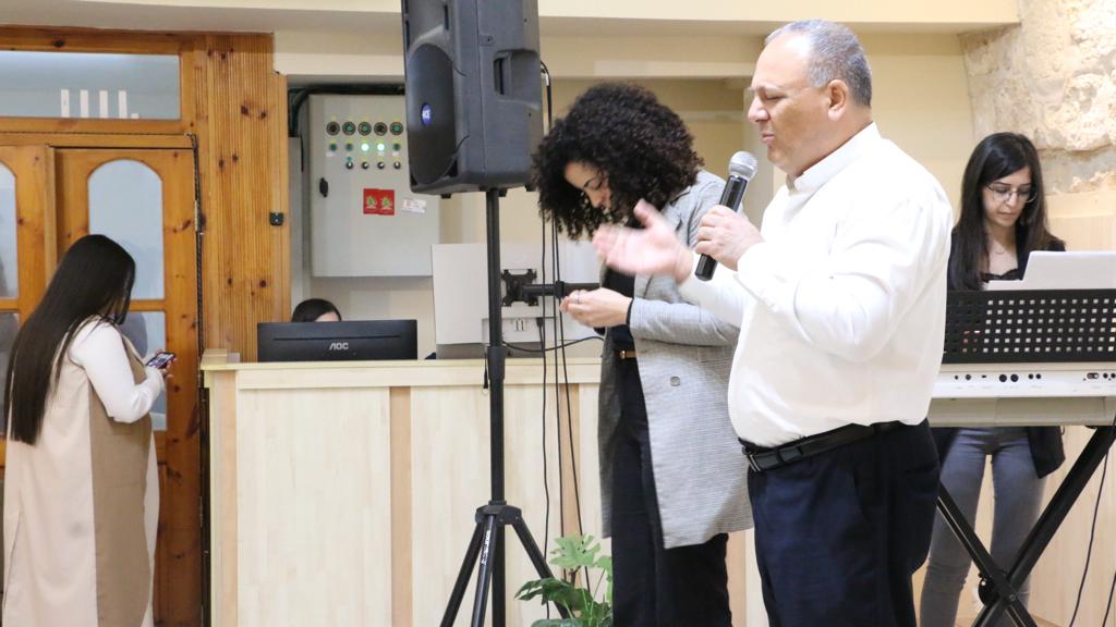 Pastor Khalid Dalleh, the Pastor of Kufur Yasif church, and also the Ministry Officer of the Baptist Association, praying over the meeting, and beside him is Luna Matar, who is the Youth Coordinator within the Baptist Association